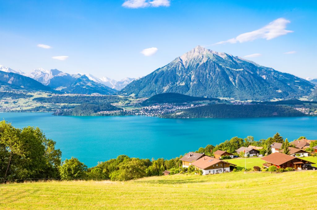 Blick von einer grünen Wiese auf einen tiefblauen See, an dessen Ufer Dörfer liegen. Im Hintergrund ragen markante, bewaldete Berge empor.